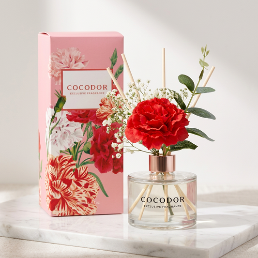 Αρωματικό χώρου Cocodor Carnation με κόκκινο γαρίφαλο, λευκά λουλούδια και ξύλινα sticks δίπλα στη ροζ συσκευασία του σε μαρμάρινη βάση - VassoHome