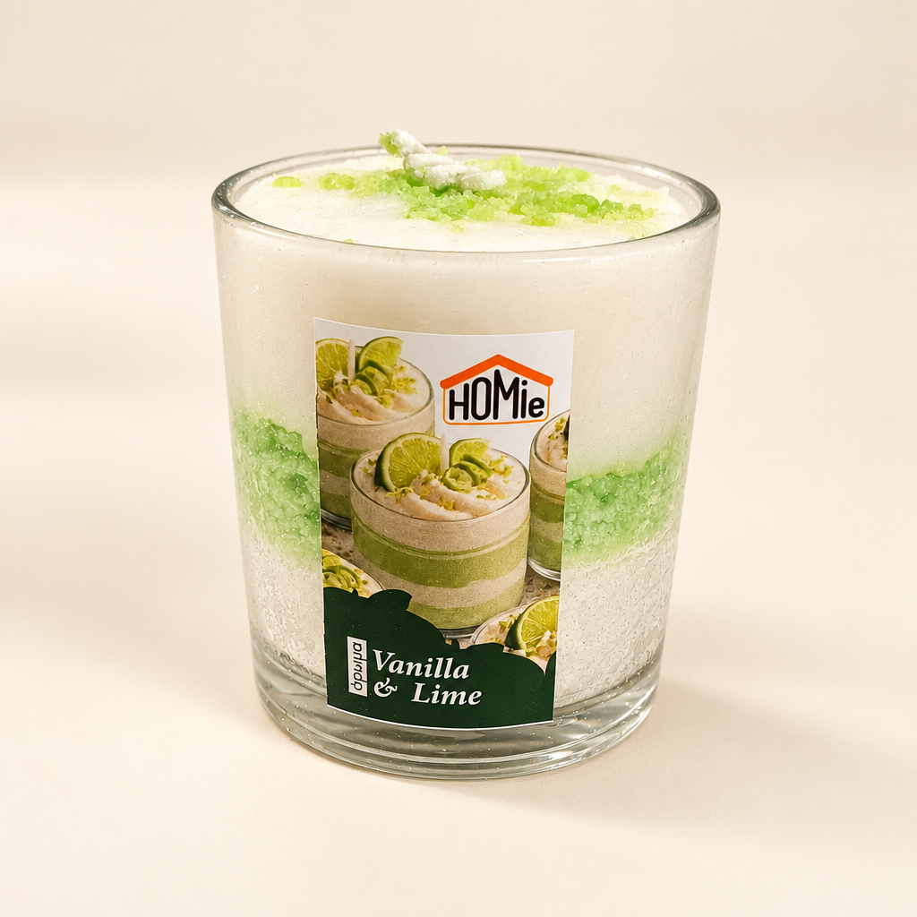 Αρωματικό κερί Vanilla & Lime σε γυάλινο ποτήρι 3/4 γωνία που αναδεικνύει την ετικέτα Homie και τα λευκά και πράσινα layers σε κρεμ φόντο - VassoHome