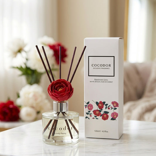 Αρωματικό χώρου Cocodor Camelia Lovely Peony 120ml με κόκκινο λουλούδι και μαύρα sticks δίπλα στη λευκή συσκευασία του σε μαρμάρινη επιφάνεια - VassoHome